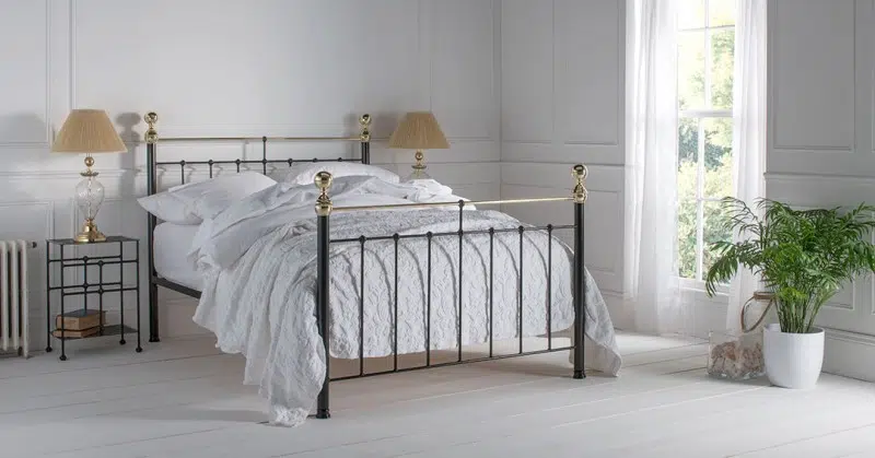 Iron and brass bed frame in a classic cottage style bedroom with white panelled walls, soft bedding, and warm natural light