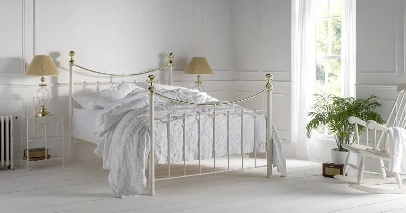 White iron and brass bed frame in a bright cottage style bedroom with panelled walls, soft white bedding, and natural light