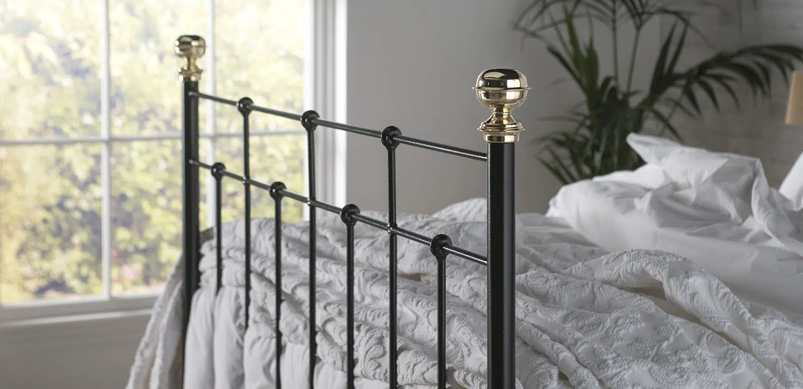 Side view of Emma bed footend showing slender black metal bars and polished brass accents in natural daylight