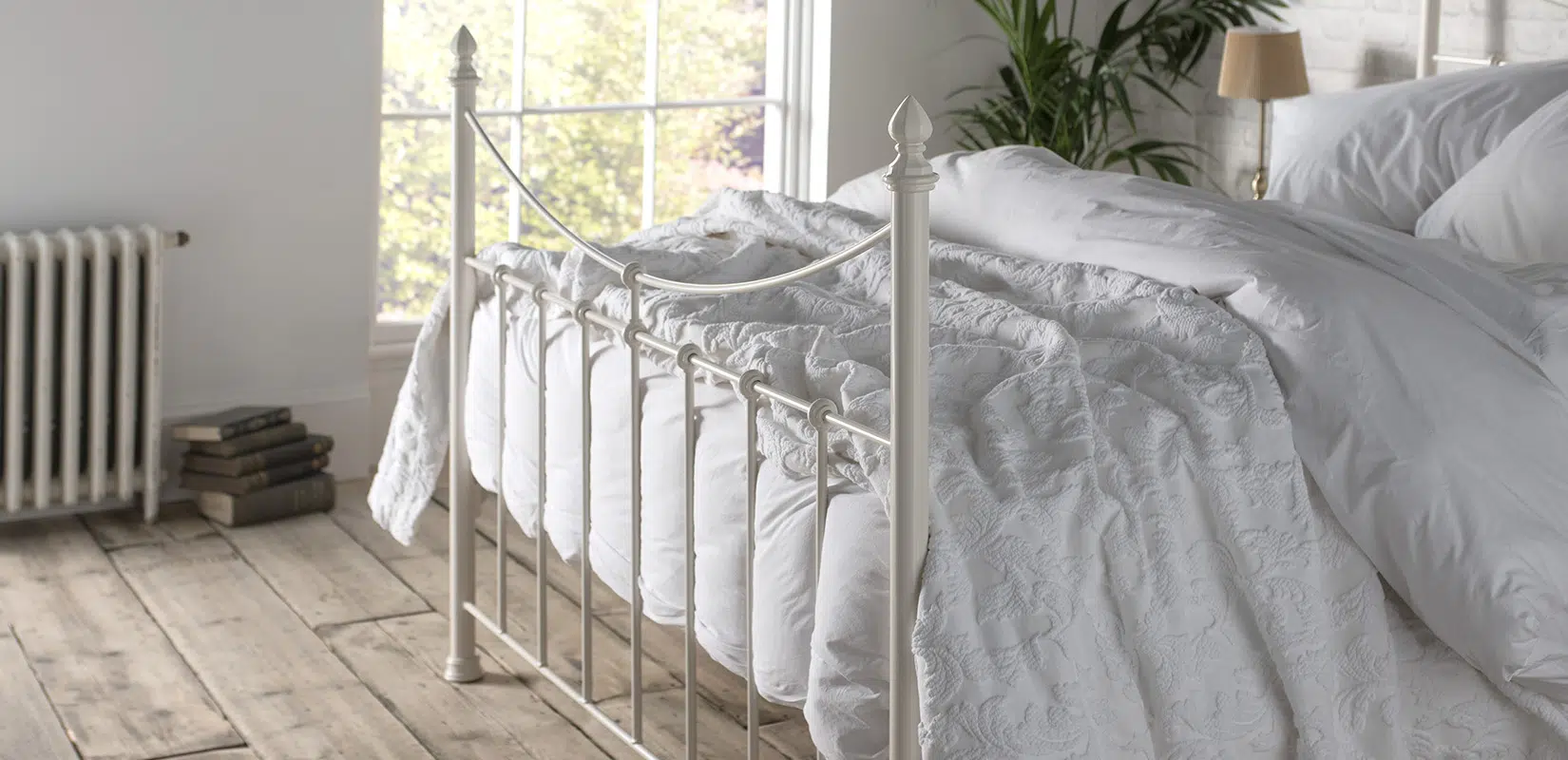 Angled view of ivory Emily iron bed showing elegant curved frame and vertical bars, placed near a large window