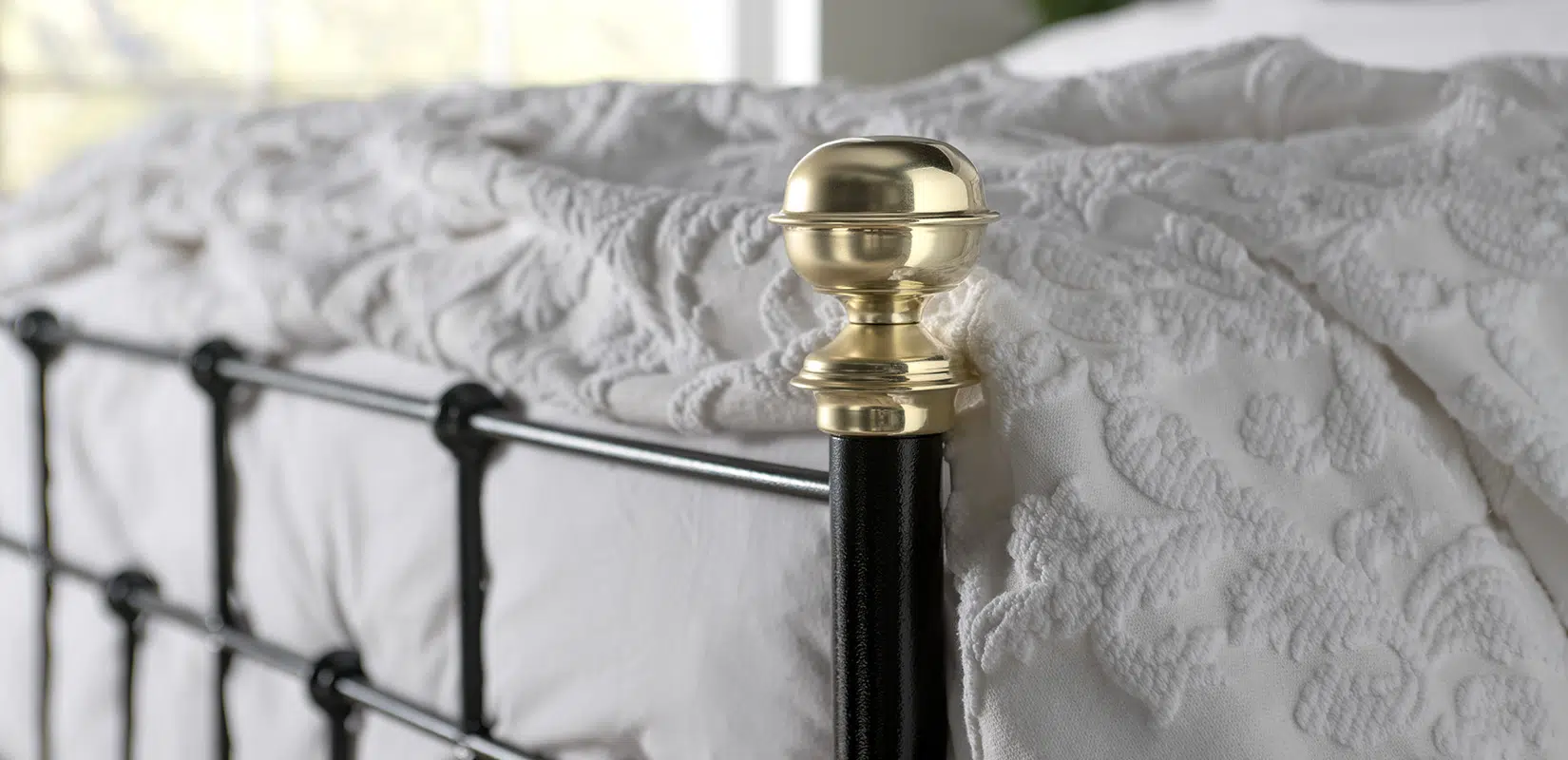 Close-up of Isaac bed’s polished brass finial on black post against soft white bedding