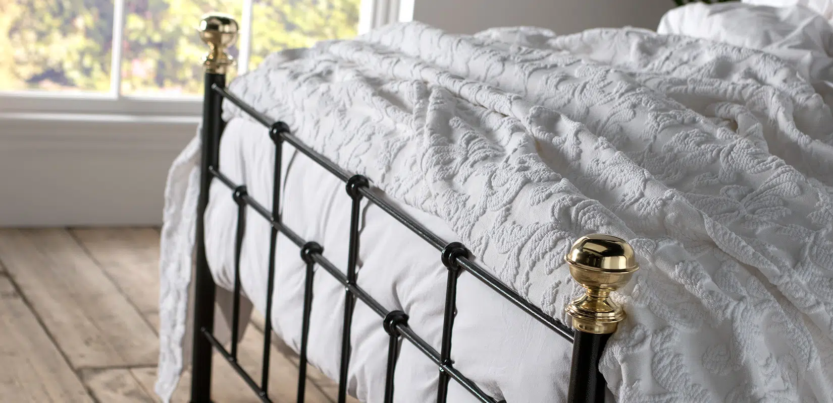 Side view of Isaac bed’s footboard with vertical bars and brass accents, styled in a sunlit bedroom