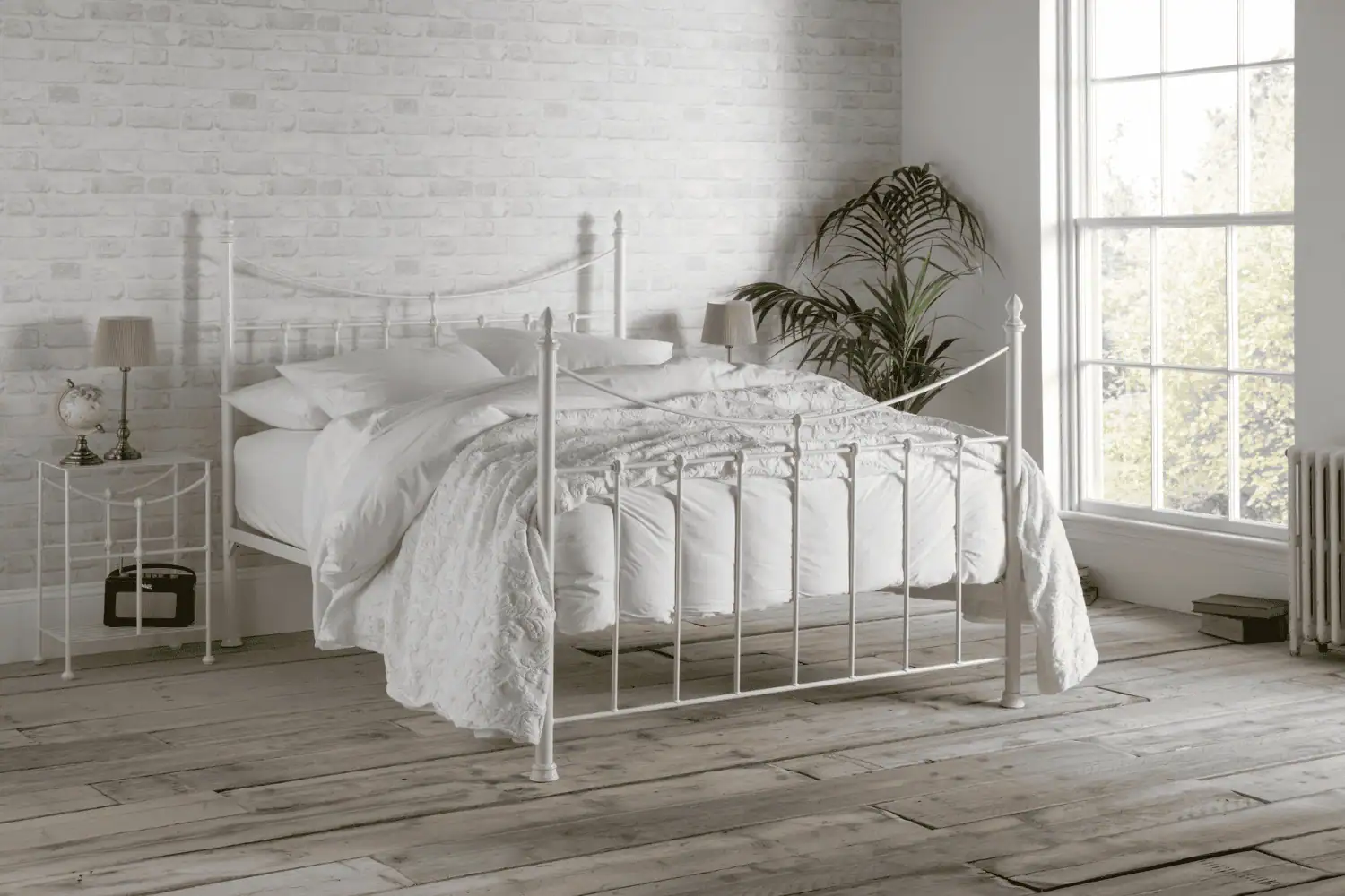 Emily iron bed in ivory finish styled with white bedding, set against a white brick wall with rustic wooden flooring
