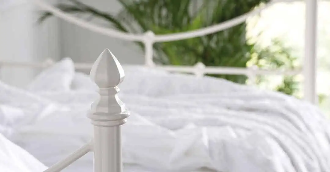 Decorative ivory finial and curved metal detailing of the Emily day bed against a soft white duvet and leafy backdrop