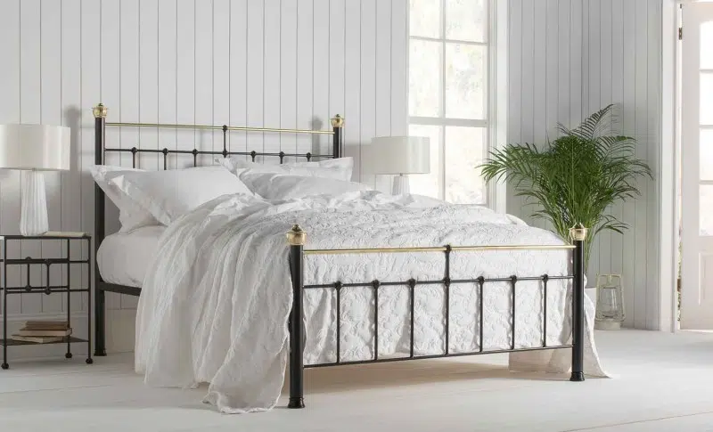 Iron and brass bed frame in a bright cottage style bedroom with white bedding, panelled walls, and soft natural textures