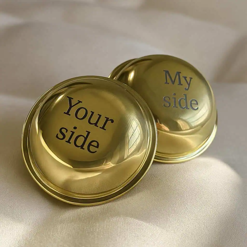 A close-up image of two polished brass finials resting on a soft cream fabric. One is engraved with the text "Your side" and the other with "My side," showcasing a playful and personalised design feature for a bed frame.