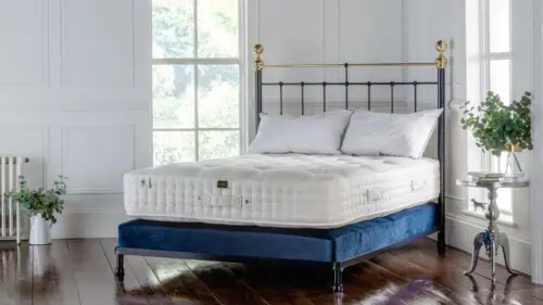 Luxurious bedroom featuring a black iron bed frame with brass finials, dressed in a plush white British wool mattress and white pillows, set atop a navy upholstered divan base in a bright, classic interior with wood flooring and large windows.