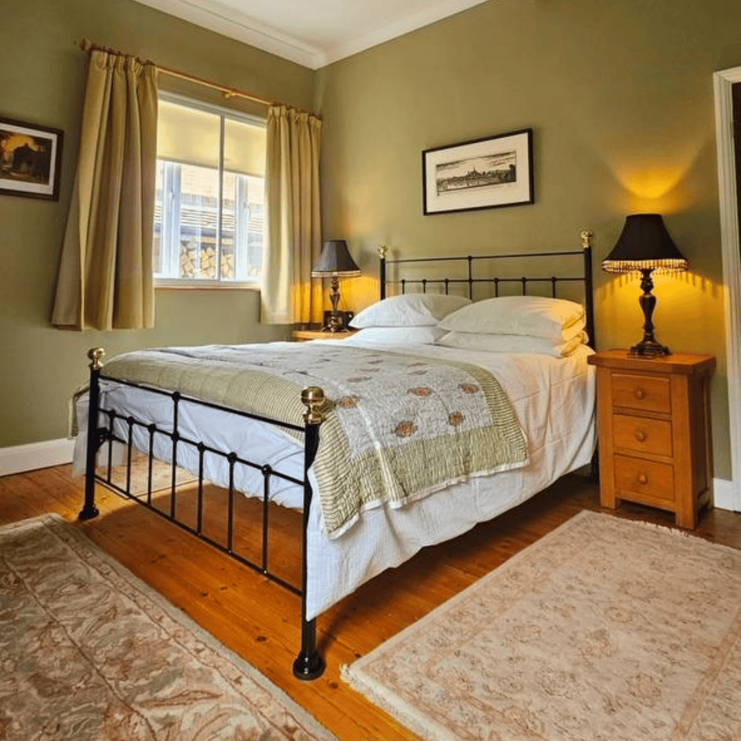A traditional black iron bed frame with white bedding in a warmly lit bedroom with wooden floors and a soft rug.