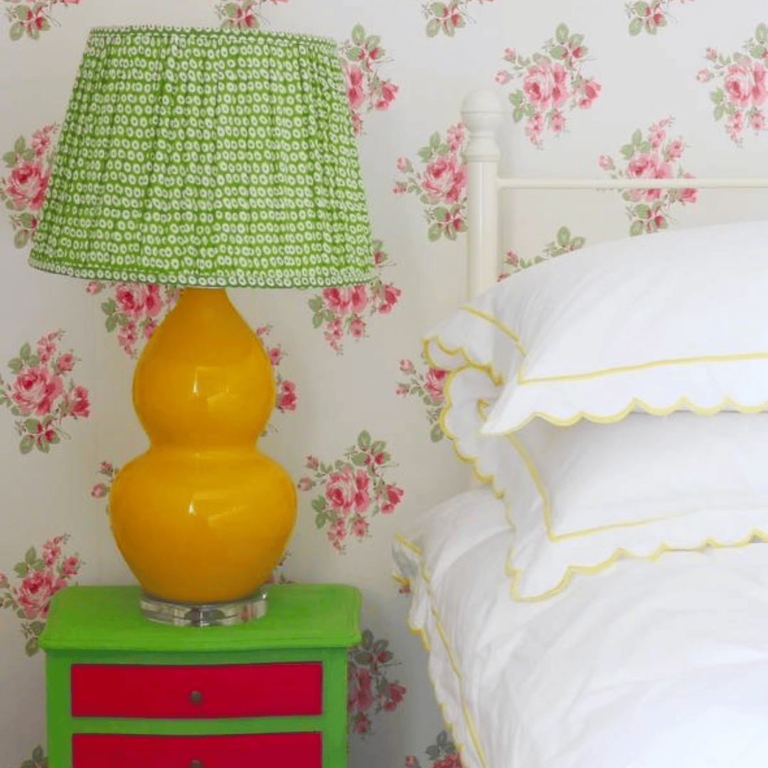 A green vintage bedside lamp with a yellow base on a small table, set against floral wallpaper and white pillows.