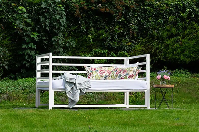 Elegant white wrought iron and brass bed with floral cushions in garden setting.